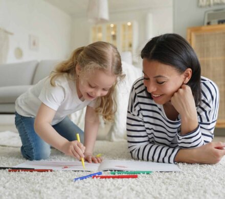 Mom, adopted child daughter draw with colorful pencils, lying on floor. Happy motherhood, adoption Smiling mother and little adopted child daughter painting together, lying floor. Mom or babysitter helps preschooler kid girl to draw with pencils. Happy motherhood, adoption concept.